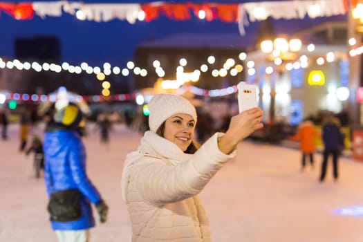 A woman on the rink is skating and taking selfie on smartphone. New Years Eve and Christmas. Fairy lights. Ice and snow mood concept. Winter