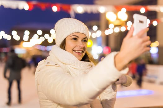 Christmas, winter, technology and leisure concept - happy young woman taking picture with smartphone on ice skating rink.