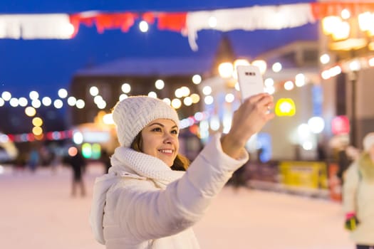 A woman on the rink is skating and taking selfie on smartphone. New Years Eve and Christmas. Fairy lights. Ice and snow mood concept. Winter