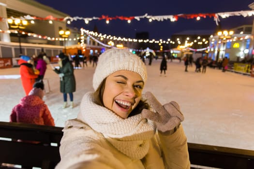 A woman on the rink is skating and taking selfie on smartphone. New Years Eve and Christmas. Fairy lights. Ice and snow mood concept. Winter