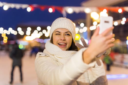 A woman on the rink is skating and taking selfie on smartphone. New Years Eve and Christmas. Fairy lights. Ice and snow mood concept. Winter