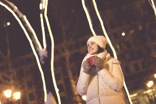 Girl standing over winter christmas city background snow snow drifts, stands warm jacket hat, holds cup tea with tea drinks.