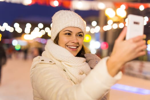 Christmas, winter, technology and leisure concept - happy young woman taking picture with smartphone on ice skating rink.