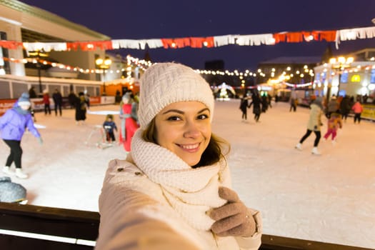 A woman on the rink is skating and taking selfie on smartphone. New Years Eve and Christmas. Fairy lights. Ice and snow mood concept. Winter