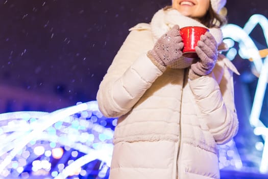 Girl standing over winter christmas city background snow snow drifts, stands warm jacket hat, holds cup tea with tea drinks.