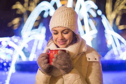 Girl standing over winter christmas city background snow snow drifts, stands warm jacket hat, holds cup tea with tea drinks.