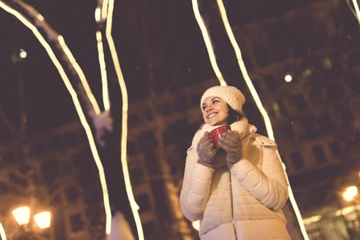 Girl standing over winter christmas city background snow snow drifts, stands warm jacket hat, holds cup tea with tea drinks.