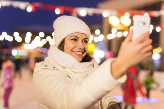 Christmas, winter, technology and leisure concept - happy young woman taking picture with smartphone on ice skating rink.