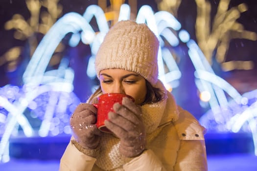 Christmas holidays. Beautiful smiling woman in warm clothing with cup of hot coffee outdoor in winter