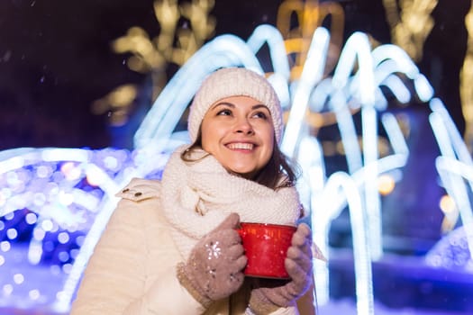 Christmas holidays. Beautiful smiling woman in warm clothing with cup of hot coffee outdoor in winter