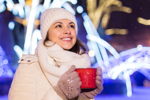 Girl standing over winter christmas city background snow snow drifts, stands warm jacket hat, holds cup tea with tea drinks.