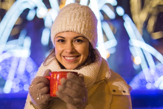 Girl standing over winter christmas city background snow snow drifts, stands warm jacket hat, holds cup tea with tea drinks.
