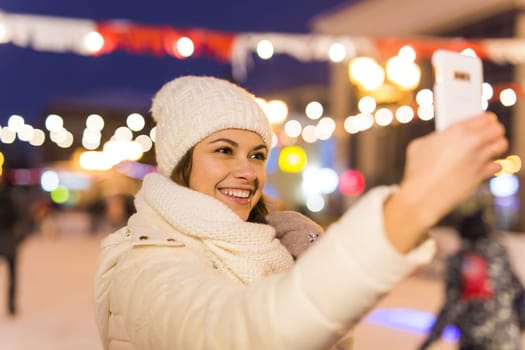 A woman on the rink is skating and taking selfie on smartphone. New Years Eve and Christmas. Fairy lights. Ice and snow mood concept. Winter
