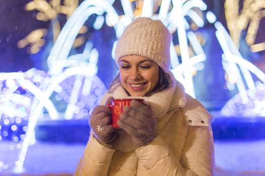 Girl standing over winter christmas city background snow snow drifts, stands warm jacket hat, holds cup tea with tea drinks.