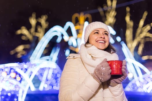 Christmas holidays. Beautiful smiling woman in warm clothing with cup of hot coffee outdoor in winter