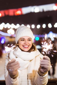 Young beautiful woman in knitted hat and scarf standing in the city with bengal light, sparkler. Concept celebration and christmas