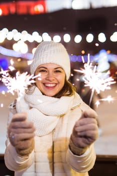 young woman wearing winter knitted clothes holding sparkler outdoors over snow background. Christmas holidays.