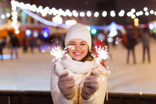 Young beautiful woman in knitted hat and scarf standing in the city with bengal light, sparkler. Concept celebration and christmas