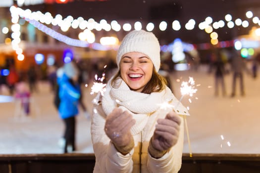 young woman wearing winter knitted clothes holding sparkler outdoors over snow background. Christmas holidays.