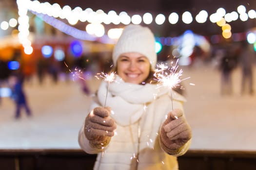 young woman wearing winter knitted clothes holding sparkler outdoors over snow background. Christmas holidays.