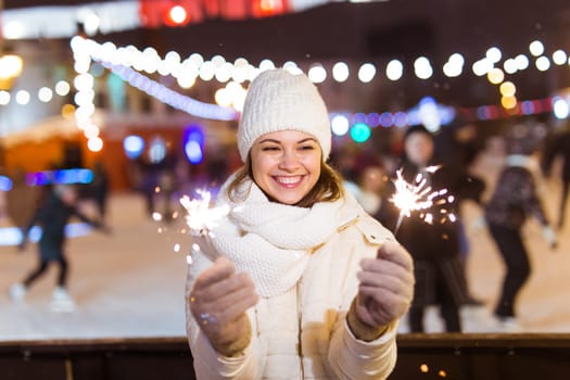 young woman wearing winter knitted clothes holding sparkler outdoors over snow background. Christmas holidays.