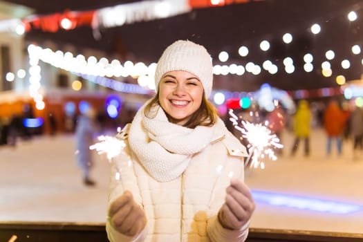 young woman wearing winter knitted clothes holding sparkler outdoors over snow background. Christmas holidays.