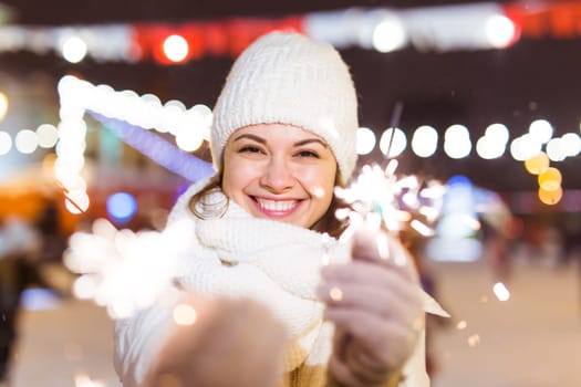 Young beautiful woman in knitted hat and scarf standing in the city with bengal light, sparkler. Concept celebration and christmas