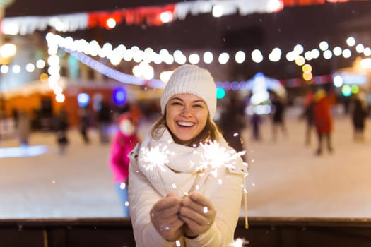 young woman wearing winter knitted clothes holding sparkler outdoors over snow background. Christmas holidays.