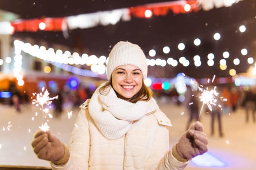 young woman wearing winter knitted clothes holding sparkler outdoors over snow background. Christmas holidays.