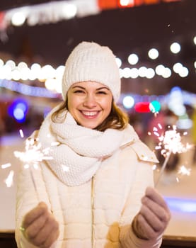 Young beautiful woman in knitted hat and scarf standing in the city with bengal light, sparkler. Concept celebration and christmas