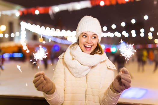 Young beautiful woman in knitted hat and scarf standing in the city with bengal light, sparkler. Concept celebration and christmas