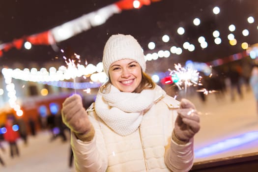 Young beautiful woman in knitted hat and scarf standing in the city with bengal light, sparkler. Concept celebration and christmas