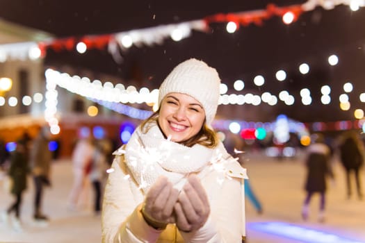 Young beautiful woman in knitted hat and scarf standing in the city with bengal light, sparkler. Concept celebration and christmas