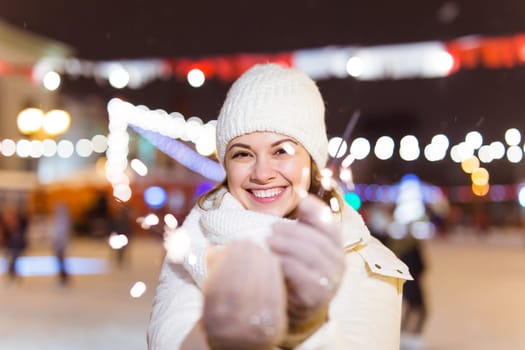 young woman wearing winter knitted clothes holding sparkler outdoors over snow background. Christmas holidays.