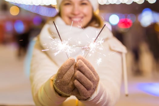 young woman wearing winter knitted clothes holding sparkler outdoors over snow background. Christmas holidays.