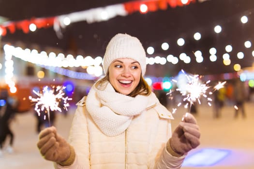 young woman wearing winter knitted clothes holding sparkler outdoors over snow background. Christmas holidays.