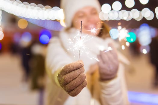 young woman wearing winter knitted clothes holding sparkler outdoors over snow background. Christmas holidays.