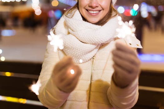 young woman wearing winter knitted clothes holding sparkler outdoors over snow background. Christmas holidays.
