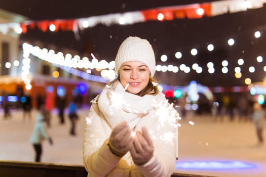 young woman wearing winter knitted clothes holding sparkler outdoors over snow background. Christmas holidays.