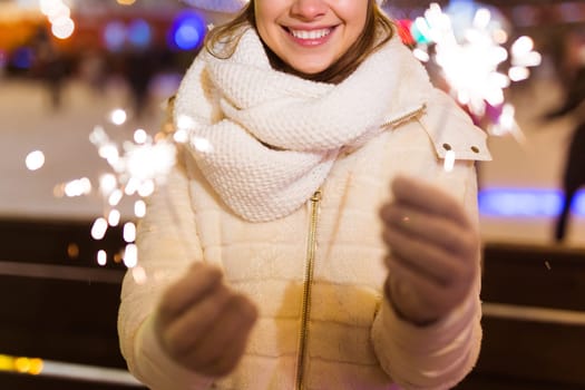 young woman wearing winter knitted clothes holding sparkler outdoors over snow background. Christmas holidays.