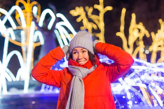 Girl having fun christmas decoration lights street. Young happy smiling woman wearing stylish knitted scarf and jacket outdoors. Model laughing. Winter wonderland city scene, New Year party.
