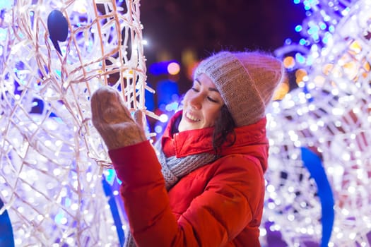 Girl having fun christmas decoration lights street. Young happy smiling woman wearing stylish knitted scarf and jacket outdoors. Model laughing. Winter wonderland city scene, New Year party.