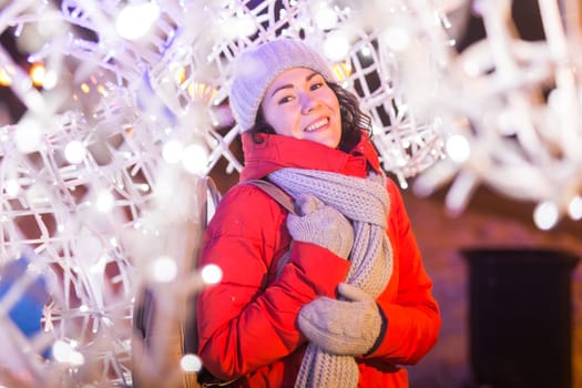 Girl having fun christmas decoration lights street. Young happy smiling woman wearing stylish knitted scarf and jacket outdoors. Model laughing. Winter wonderland city scene, New Year party.