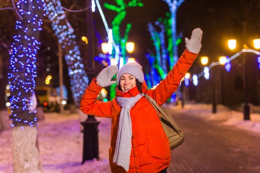 Girl having fun christmas decoration lights street. Young happy smiling woman wearing stylish knitted scarf and jacket outdoors. Model laughing. Winter wonderland city scene, New Year party.