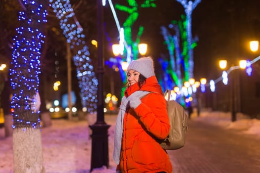 Girl having fun christmas decoration lights street. Young happy smiling woman wearing stylish knitted scarf and jacket outdoors. Model laughing. Winter wonderland city scene, New Year party.