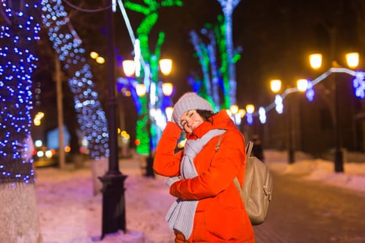 Girl having fun christmas decoration lights street. Young happy smiling woman wearing stylish knitted scarf and jacket outdoors. Model laughing. Winter wonderland city scene, New Year party.