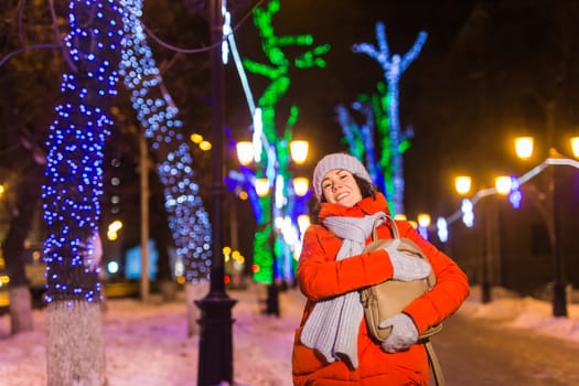 Girl having fun christmas decoration lights street. Young happy smiling woman wearing stylish knitted scarf and jacket outdoors. Model laughing. Winter wonderland city scene, New Year party.