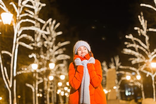 Girl having fun christmas decoration lights street. Young happy smiling woman wearing stylish knitted scarf and jacket outdoors. Model laughing. Winter wonderland city scene, New Year party.