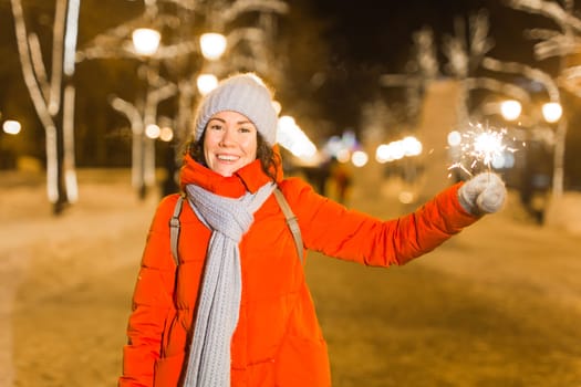 young woman wearing winter knitted clothes holding sparkler outdoors over snow background. Christmas holidays.