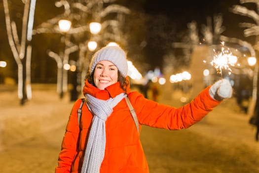 young woman wearing winter knitted clothes holding sparkler outdoors over snow background. Christmas holidays.